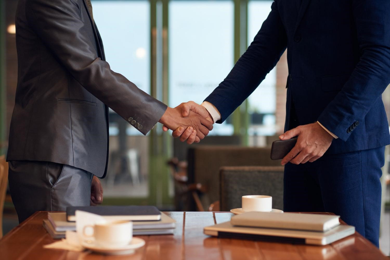 Two people in suits shake hands across a table with notebooks and coffee cups in a modern office setting. Business-like mood.