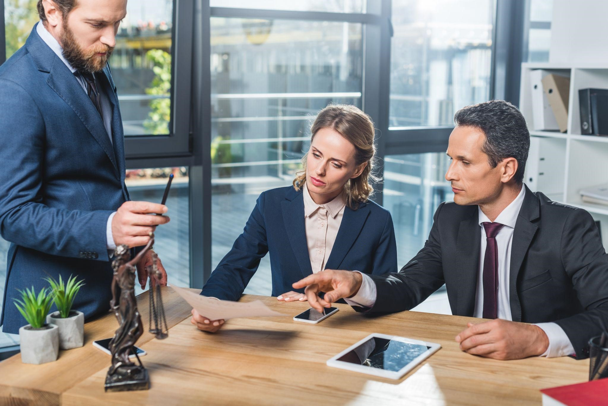 Three professionals in a modern office discuss a document. A Justice statue and plants are on the table. The mood is focused.