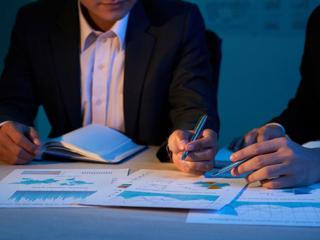 Businessmen in suits analyze graphs and charts under blue lighting, pointing with pens. Papers are scattered on a wooden table.