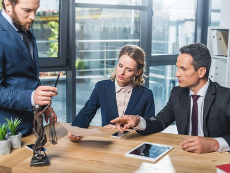 Three professionals in a modern office discuss a document. A Justice statue and plants are on the table. The mood is focused.