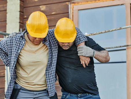 A construction worker with a hand on her lower back is helped to walk by a colleague at a site, representing a workplace injury and workers' comp claim.