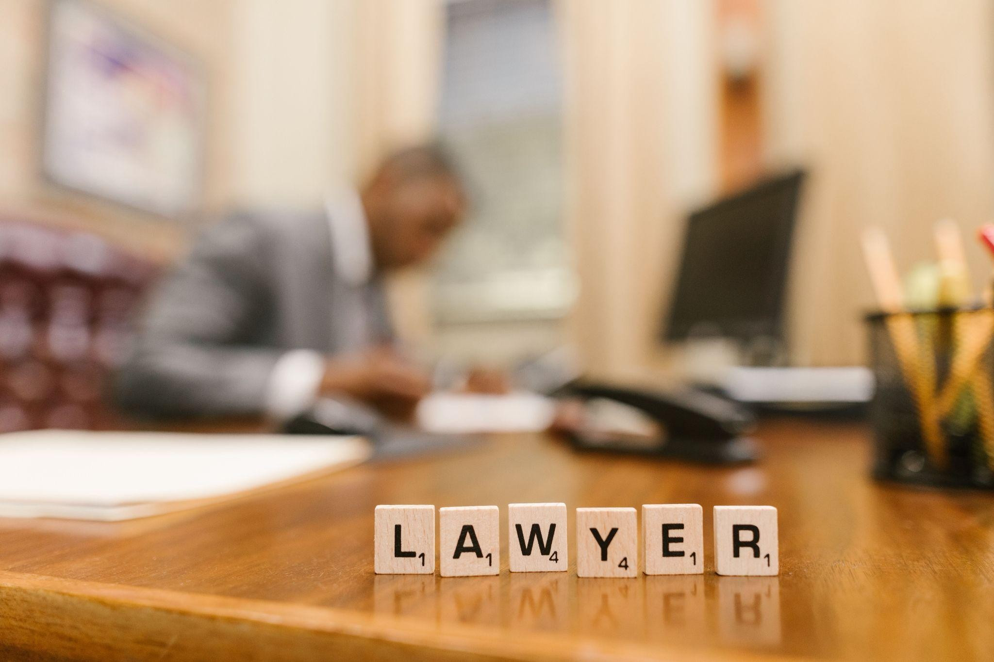 Scrabble tiles spelling LAWYER on a wooden desk, with a blurred lawyer working in an office behind them.