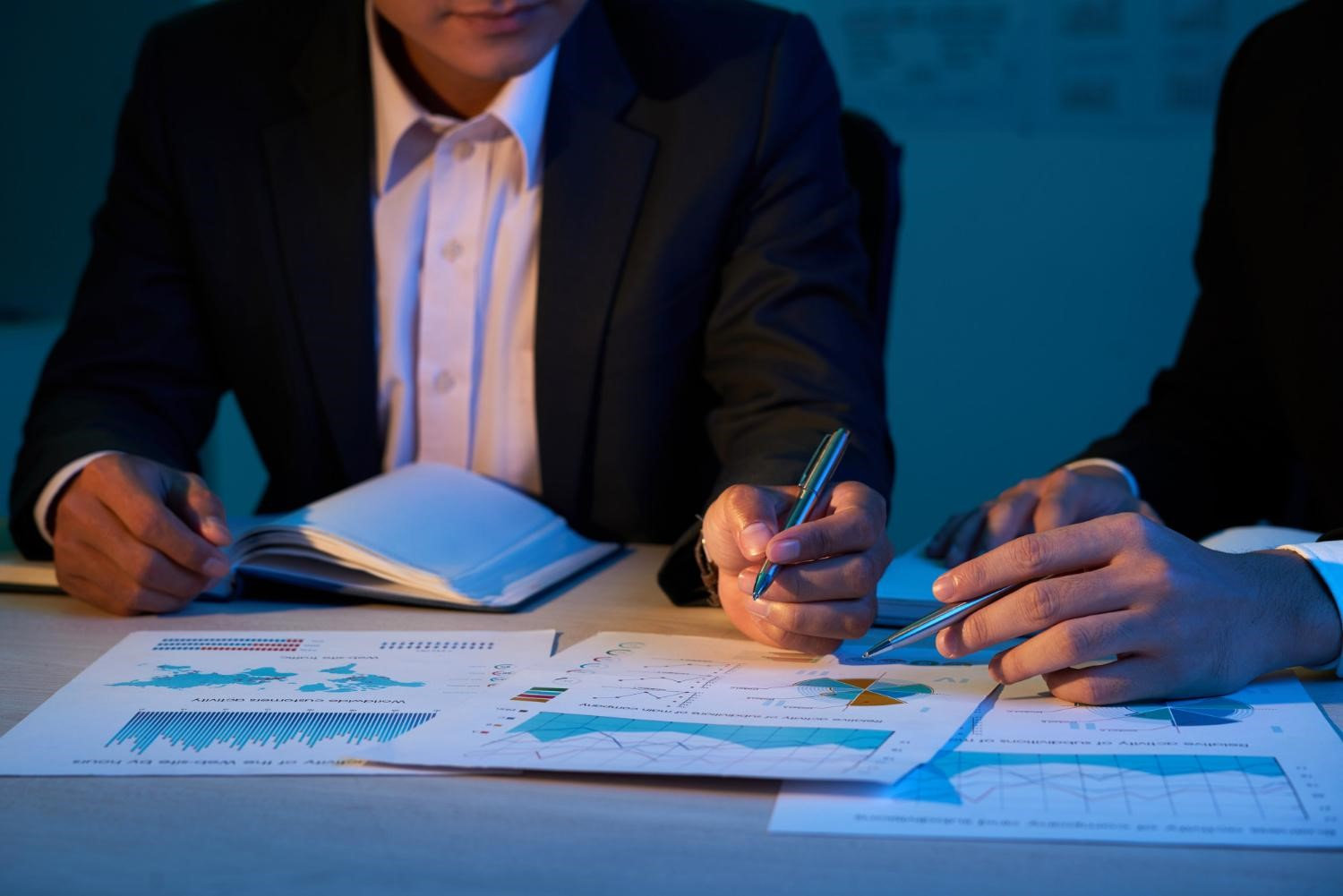 Businessmen in suits analyze graphs and charts under blue lighting, pointing with pens. Papers are scattered on a wooden table.