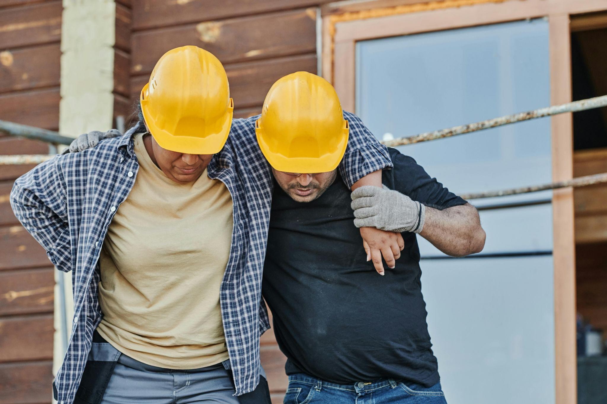 A construction worker with a hand on her lower back is helped to walk by a colleague at a site, representing a workplace injury and workers' comp claim.