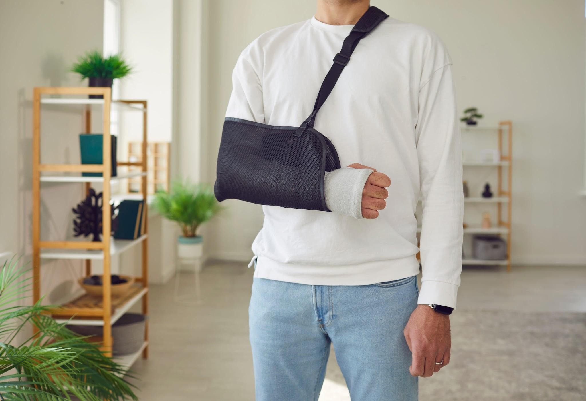 Person wearing a white sweatshirt with an arm in a sling and a bandaged wrist, standing at home while recovering from a workplace