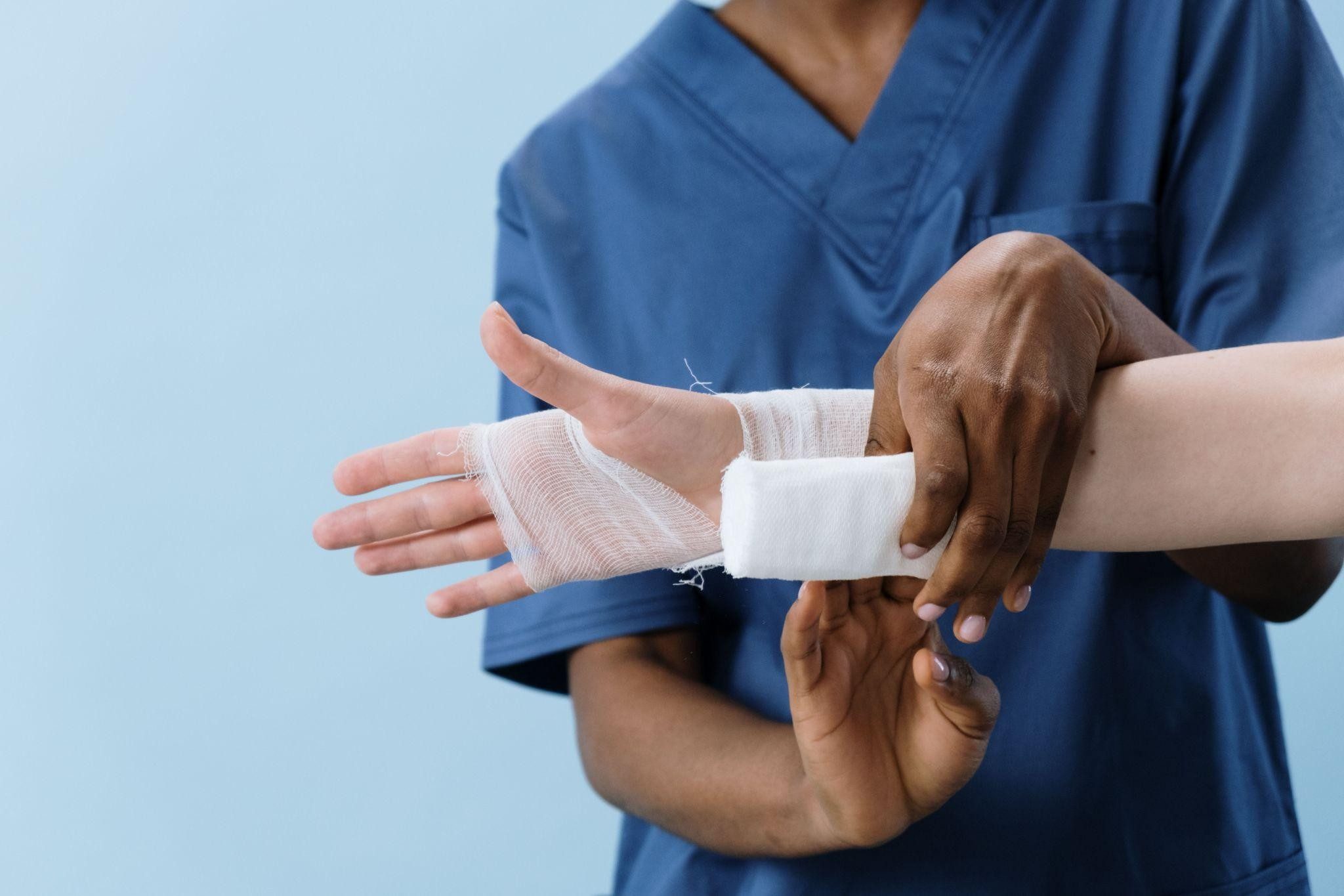 Nurse in blue scrubs wraps a bandaged wrist and hand against a light blue background, suggesting careful medical treatment