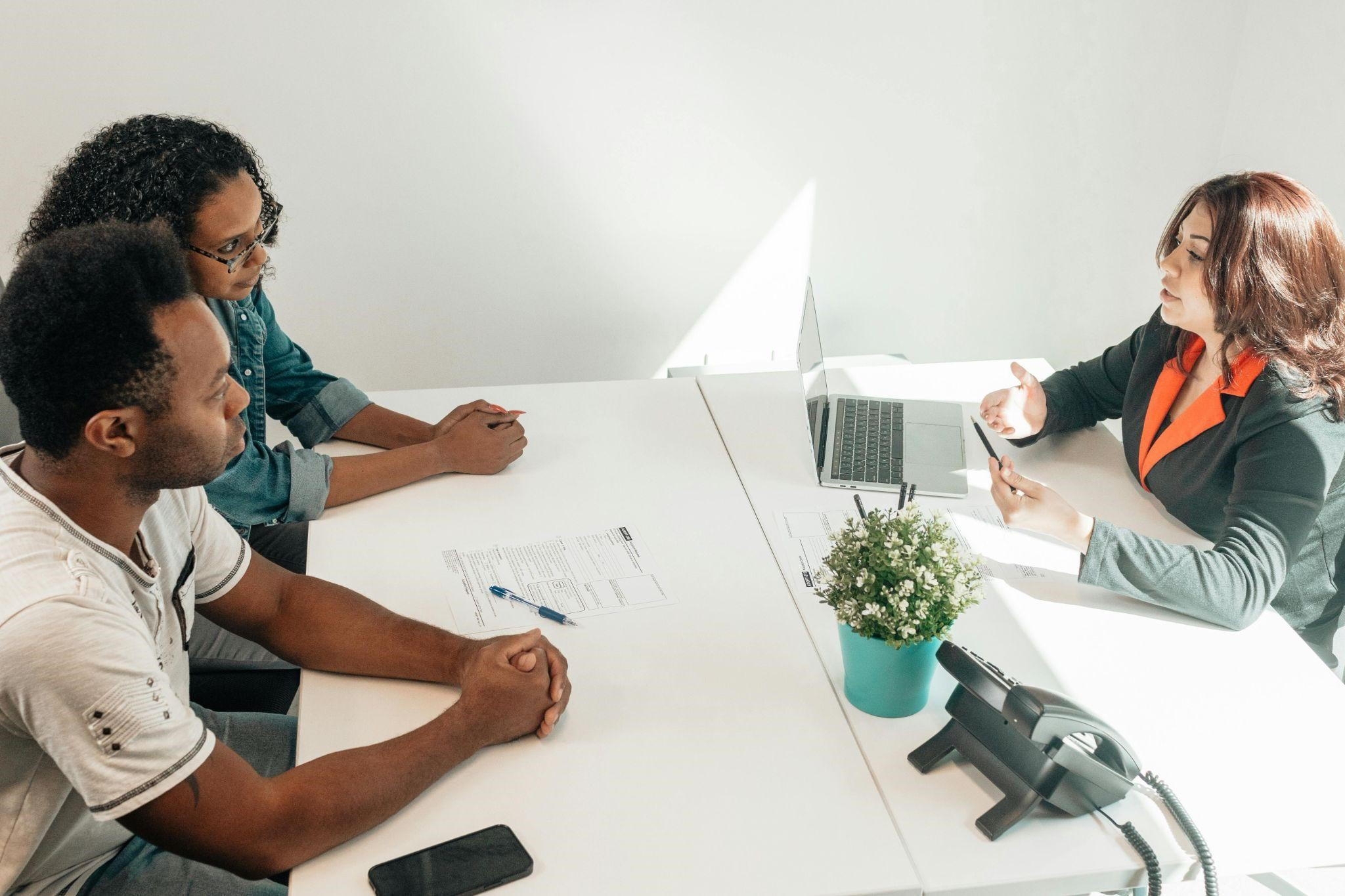 Two people sit across from a woman at a desk in a meeting. A laptop, documents, and a potted plant are on the white table.