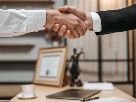 Two people shaking hands in an office setting. A laptop, framed certificate, and cup of coffee are on the desk. Professional atmosphere.