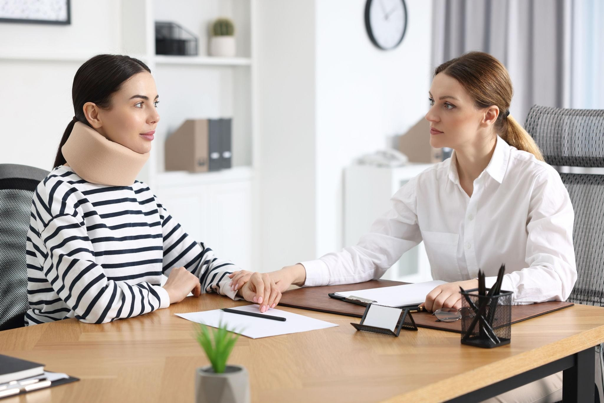 Two women in an office, one wearing a neck brace and striped shirt, the other in a white blouse, hold hands across a desk. Calm and supportive mood.