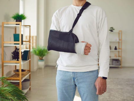 Person wearing a white sweatshirt with an arm in a sling and a bandaged wrist, standing at home while recovering from a workplace